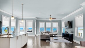 A spacious living area with a large window overlooking the sea, a kitchen with white cabinetry, and a ceiling fan.