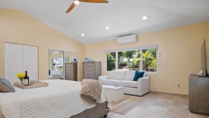 A bedroom with a bed, a sofa, a desk, and a wardrobe. There is a window with a view of greenery outside. - Exquisite Courtyard Villa on St. Thomas East End / Ocean Views + Modern Comfort (St Thomas)