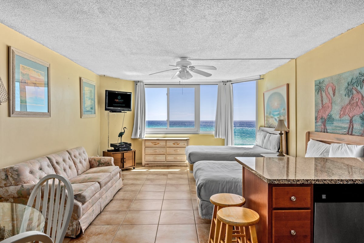 A room with a bed, a sofa, a dining table, and a TV. There is a window with a view of the ocean.