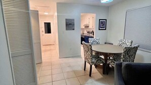 A spacious living area with a dining table and chairs, a kitchen in the background, and a hallway leading to another room.