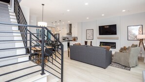 Un salon moderne avec un canapé gris, un coin repas avec une table et des chaises, une cuisine avec des meubles blancs et un escalier avec des rampes noires.