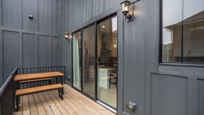 Une terrasse en bois avec un banc, une porte-fenêtre coulissante donnant sur la cuisine et un luminaire fixé au mur.