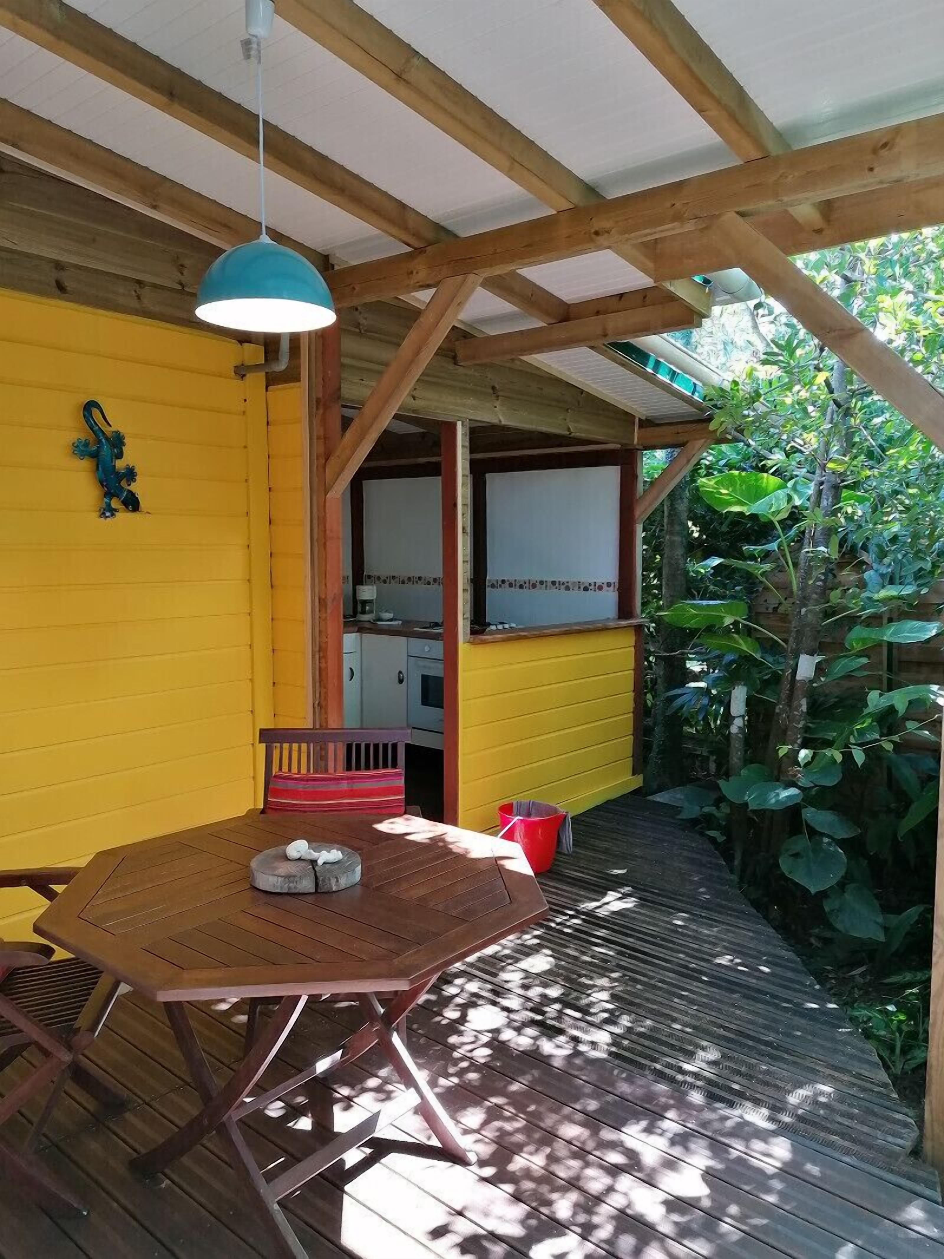 A wooden deck with a table and chairs, a hanging light fixture, and a yellow wall with a window.