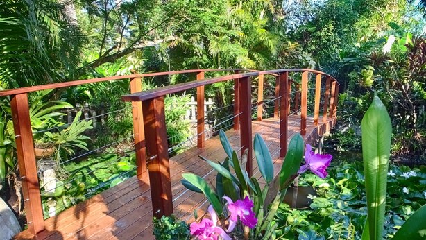 A wooden deck with a metal railing, surrounded by lush greenery and various plants. - Le Parc aux Orchidées (Pointe-Noire)