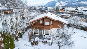 Ein zweistöckiges Holzhaus mit Balkon, umgeben von schneebedeckten Bäumen und Bergen im Hintergrund.