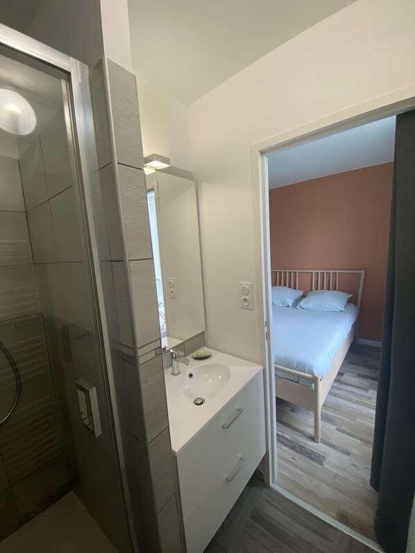 Un baño moderno con ducha, lavamanos y espejo. En la habitación contigua, hay una cama con cabecera de madera y ropa de cama blanca.