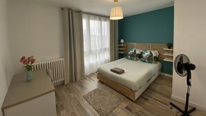 A modern bedroom with a bed, bedside table, radiator, curtains, and a standing fan. - Le Majestic (Saint-Étienne)