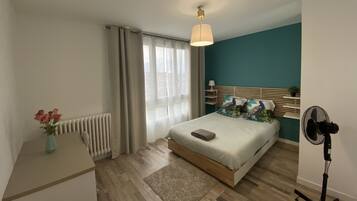 Une chambre à coucher moderne avec un lit, une table de chevet, un radiateur, des rideaux et un ventilateur sur pied.