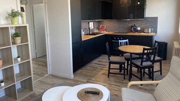 A modern kitchen with dark cabinets, a wooden countertop, and a built-in oven. There is a dining area with a round table and chairs, and a bookshelf with plants. - Le Majestic (Saint-Étienne)