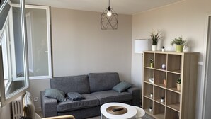 A modern living room with a grey sofa, a small white table, a wooden bookshelf, and a hanging light fixture. - Le Majestic (Saint-Étienne)