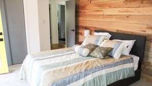 En dubbelsäng med ett randigt överkast och flera kuddar, en sänggavel i trä och en dörr som leder till ett badrum.