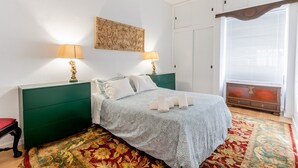 Une chambre à coucher comprenant un lit, deux tables de chevet, une commode, une fenêtre avec des stores et un tapis à motifs.
