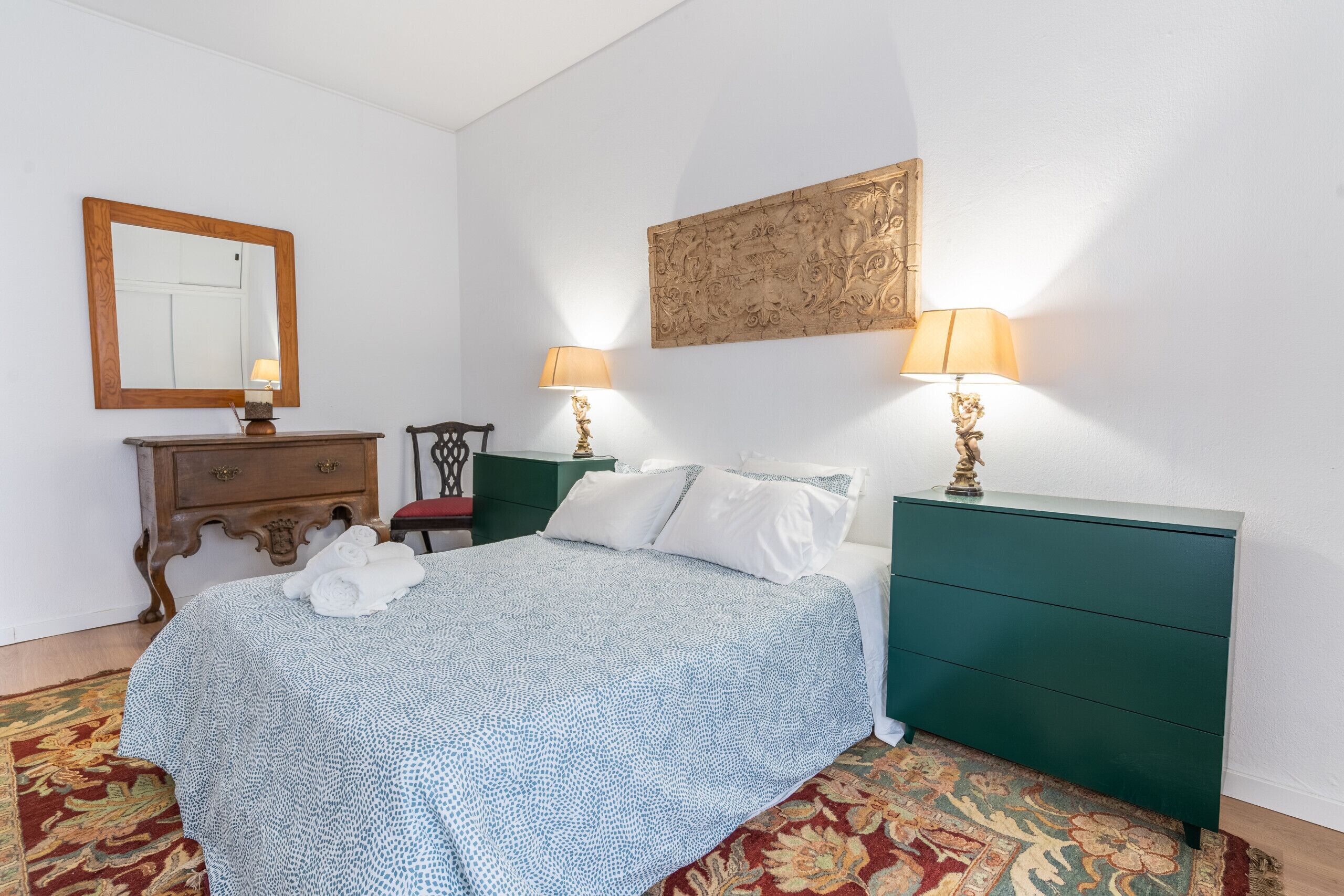 Une chambre à coucher comprenant un lit, deux tables de chevet avec des lampes, une commode, une chaise, un miroir et un élément décoratif mural.