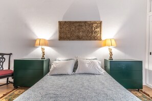 Un lit recouvert d’une couvre-lit à motifs, deux tables de chevet vertes avec des lampes, une chaise blanche et un élément décoratif mural.