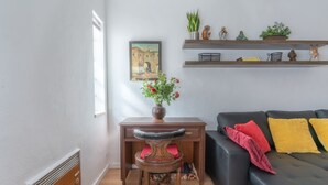 Une pièce comprenant un bureau, une chaise et un canapé noir agrémenté de coussins rouges et jaunes. On y trouve également des étagères avec des plantes et des figurines, ainsi qu’un tableau accroché au mur.
