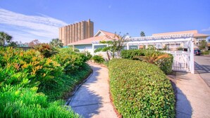 Un sendero conduce a un edificio con entrada porticada, rodeado de exuberante vegetación y una imponente estructura hotelera al fondo.