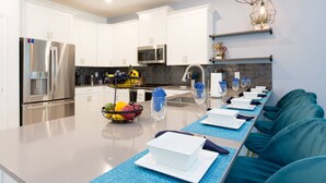 Cocina moderna con armarios blancos, electrodomésticos de acero inoxidable y una isla central con fruteros y copas de vino.