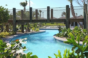Área de piscina com uma ponte de madeira, cercada por vegetação e palmeiras.