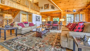 A spacious living room with a high ceiling, wooden beams, a staircase, and a large window providing natural light. - Views, Hot Tub, Fire Pit, A Mountain Masterpiece (Maggie Valley)
