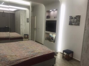 Ein Schlafzimmer mit einem großen Bett, einem Kleiderschrank, einem kleinen Hocker und einem Regal mit Dekorationen.