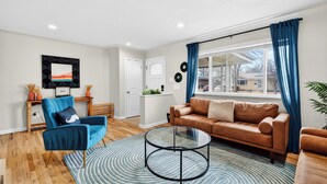 A modern living room with a brown sofa, a blue armchair, a glass coffee table, and a large window with blue curtains.