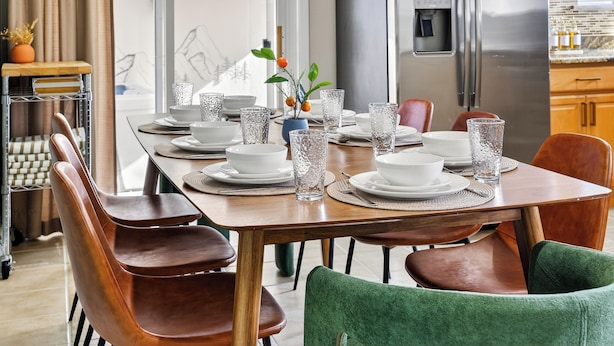 A dining area with a wooden table set for six, surrounded by leather chairs. A stainless steel refrigerator and a water dispenser are visible in the background.