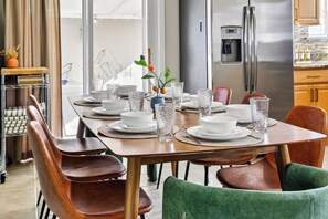 Un comedor con una mesa de madera dispuesta para seis personas, rodeada de sillas de cuero. Se observa un refrigerador de acero inoxidable y un dispensador de agua al fondo.
