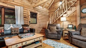 Interior de una cabaña de troncos con un sofá, una mesa de centro de madera, un sillón de cuero y una lámpara.