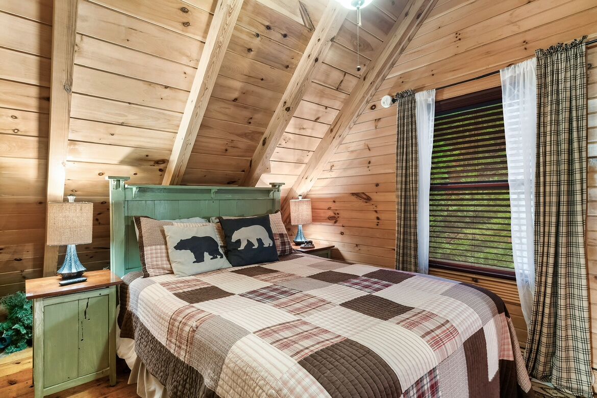 A cozy bedroom with a wooden ceiling, a green headboard, a quilted bedspread, and a window with curtains.