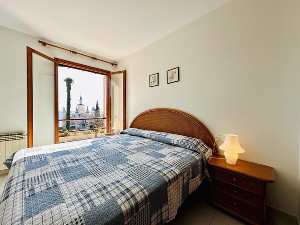 A bedroom with a large bed, a nightstand, a lamp, and a view of a castle through an open window. - Punxes CostaBravaSi - Pool, garden, beach, wifi (Sant Feliu de Guíxols)