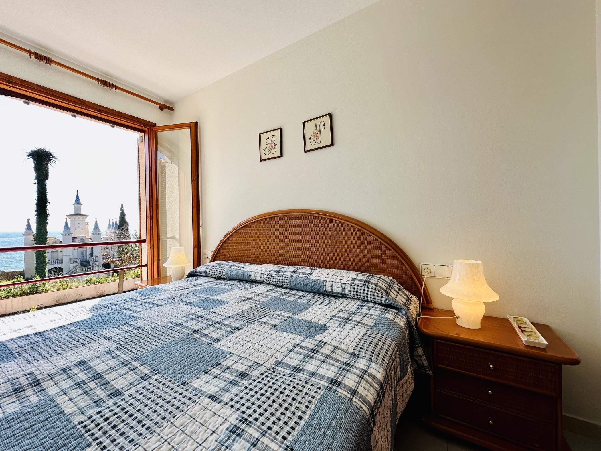 A bedroom with a large bed, a nightstand, a lamp, and a view of a castle through a window.