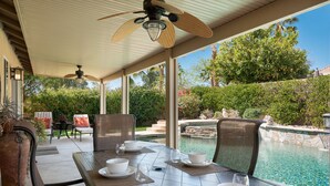 An outdoor patio with a dining table set for four, overlooking a swimming pool and lush greenery. - 5 min to Coachella - Boho Chic Oasis with Pool & Spa (Indio)