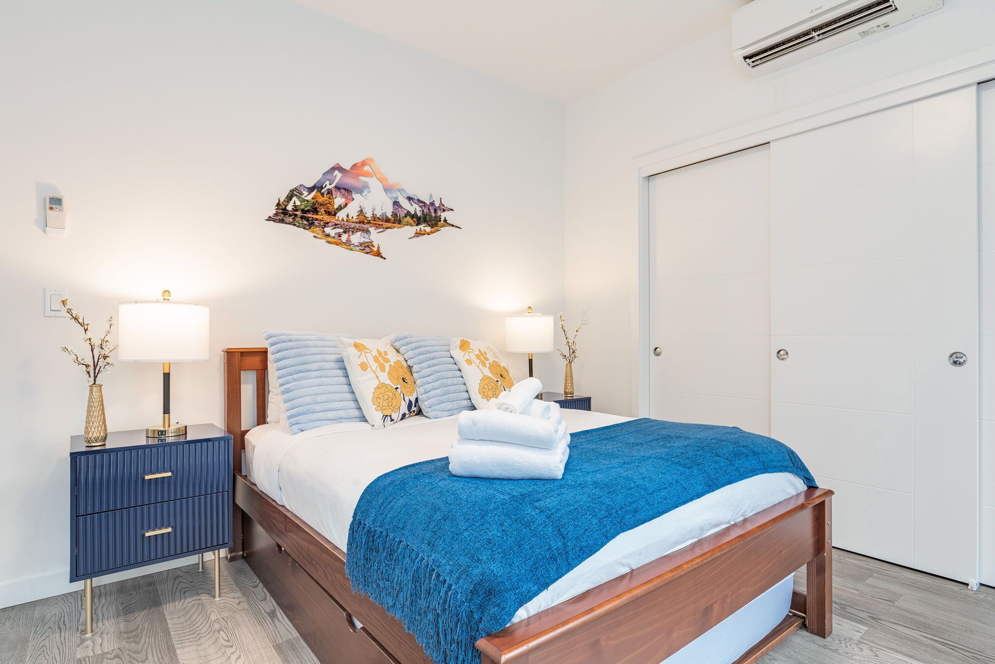 A neatly made bed with a blue blanket, two bedside tables with lamps, and a wall-mounted air conditioner.
