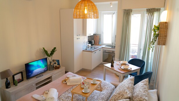 Una habitación de hotel moderna con sofá, televisor, mesa de comedor con bandeja, zona de cocina y balcón con cortinas.