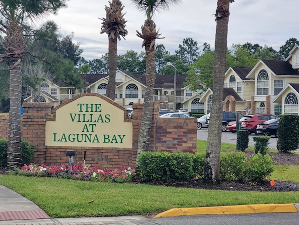 Naamplaatje voor 'The Villas at Laguna Bay' met een bakstenen voetstuk en groene letters, omringd door palmbomen en een parkeerplaats.