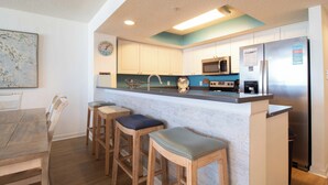 A kitchen with white cabinets, a stainless steel refrigerator, a microwave, and a sink. There are three wooden bar stools with cushioned seats. - Ocean Front Oasis with pool access~ Salty Sanctuary (Folly Beach)