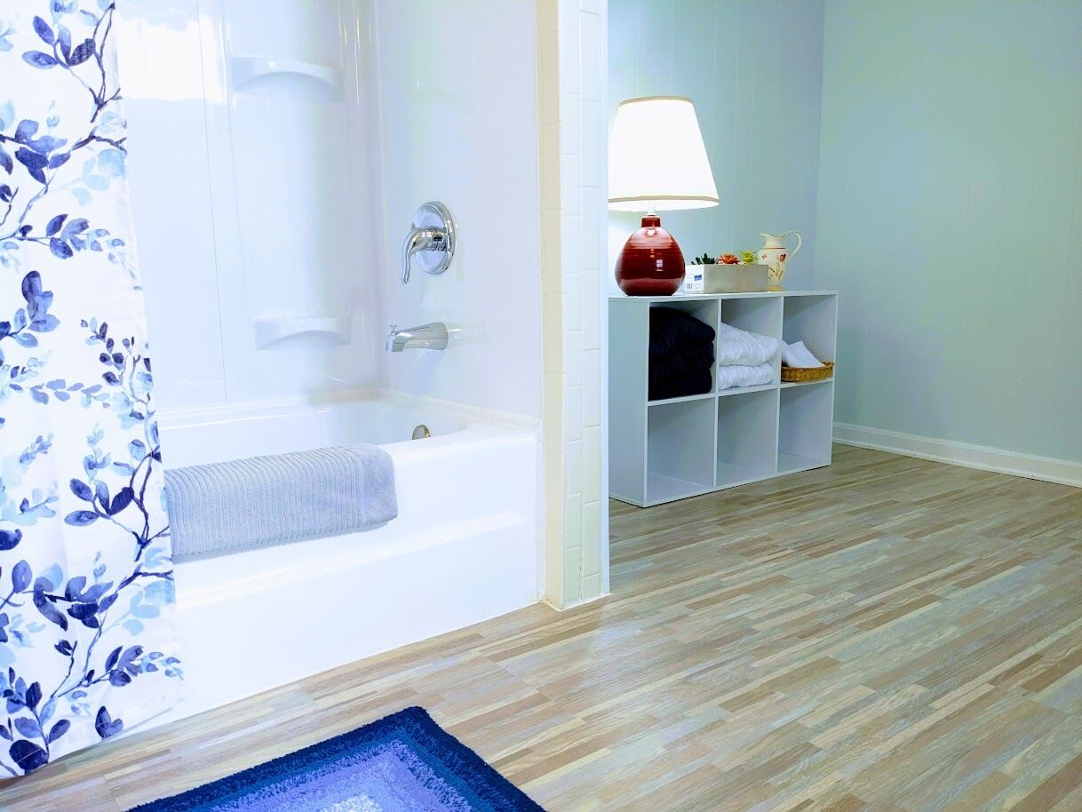 A bathroom with a bathtub, a shower, a toilet, a sink, a mirror, a shelf with toiletries, and a wooden floor.