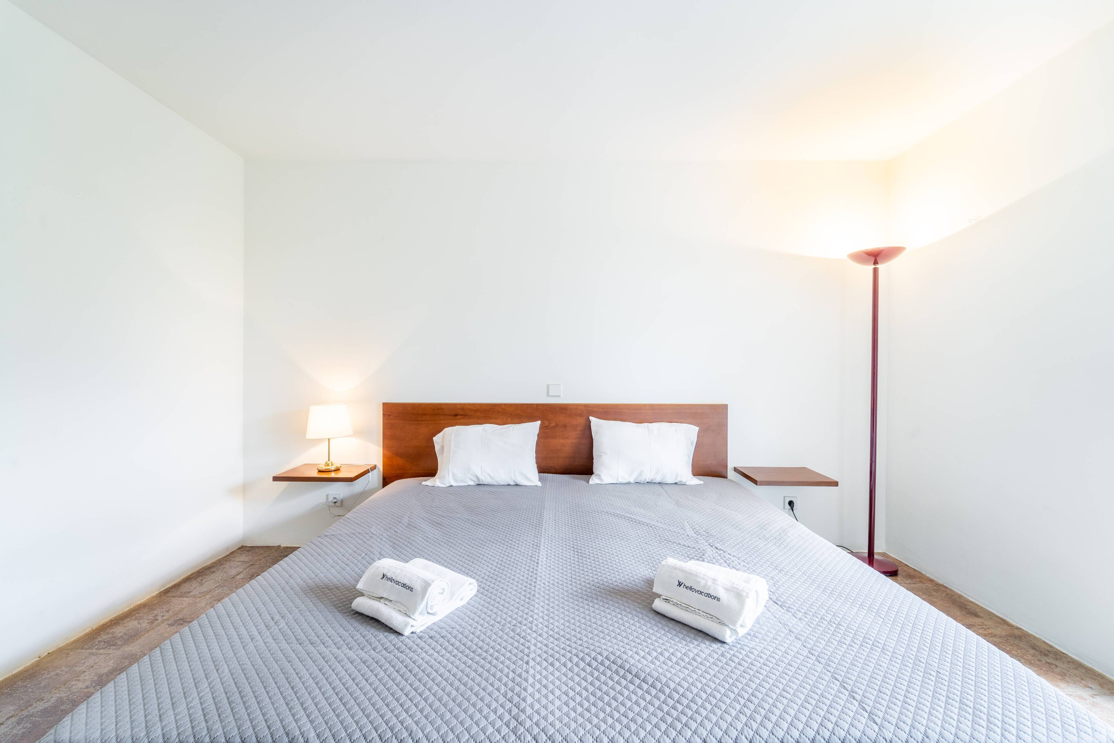 A neatly made bed with a grey quilt, two white pillows, and two rolled towels on the bed.