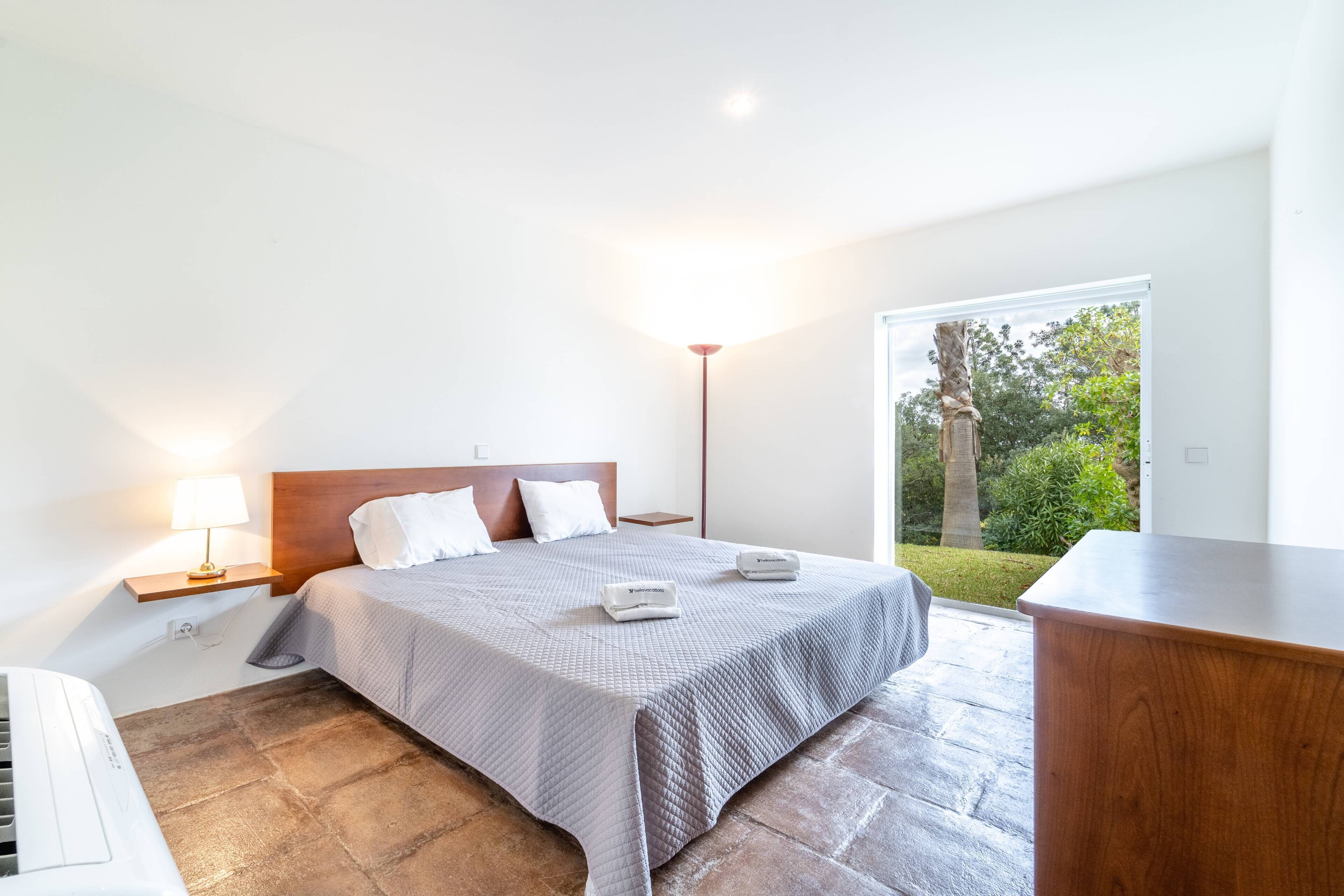 A modern bedroom with a large bed, a bedside table with a lamp, and a view of the outdoors.
