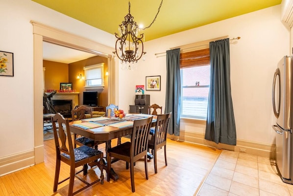 A dining area with a wooden table, chairs, and a chandelier. There is a kitchen visible in the background with a refrigerator and a window with curtains. - Beautiful Historic Home near the beach (Goderich)