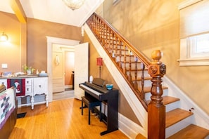 A living room with a piano, wooden staircase, and a kitchen area. - Beautiful Historic Home near the beach (Goderich)