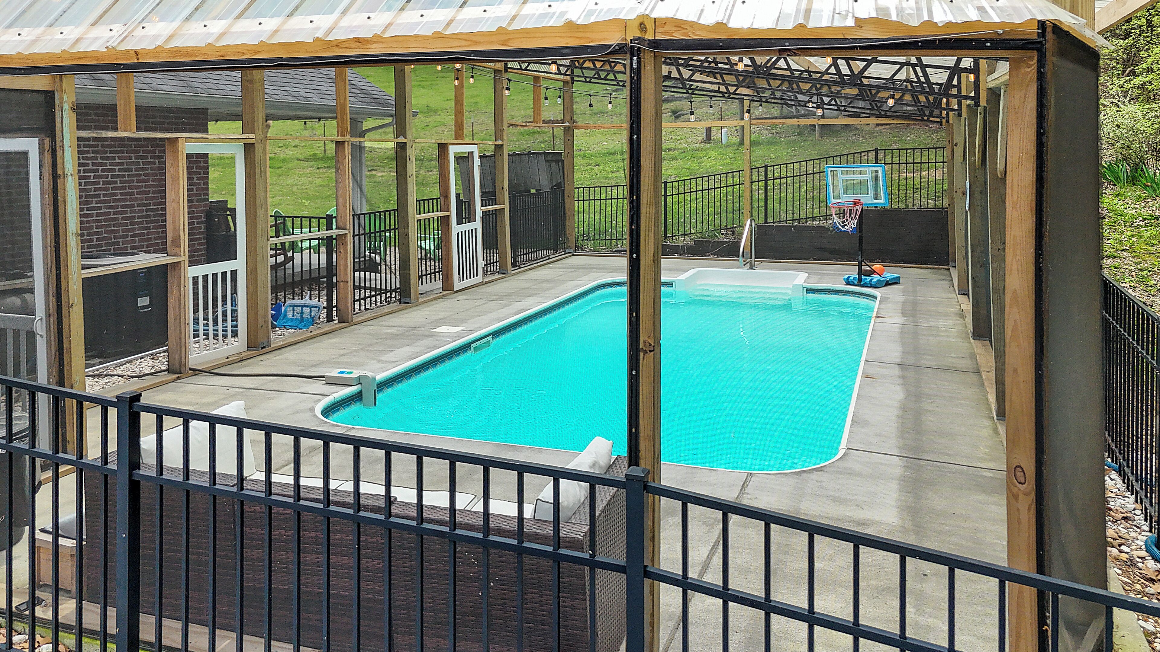 A covered swimming pool with a metal railing, a basketball hoop, and a patio area with outdoor furniture.