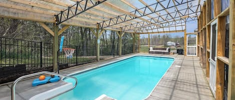 A covered swimming pool with a metal roof and wooden beams, surrounded by a black metal fence.