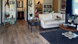 A spacious living room with a white leather sofa, a chandelier, wooden flooring, and a large window with blue curtains. - THE LOFT an UPSCALE RUSTIC 2 Bedroom 2 Washroom Private Guest House on 16acres. (Tottenham)