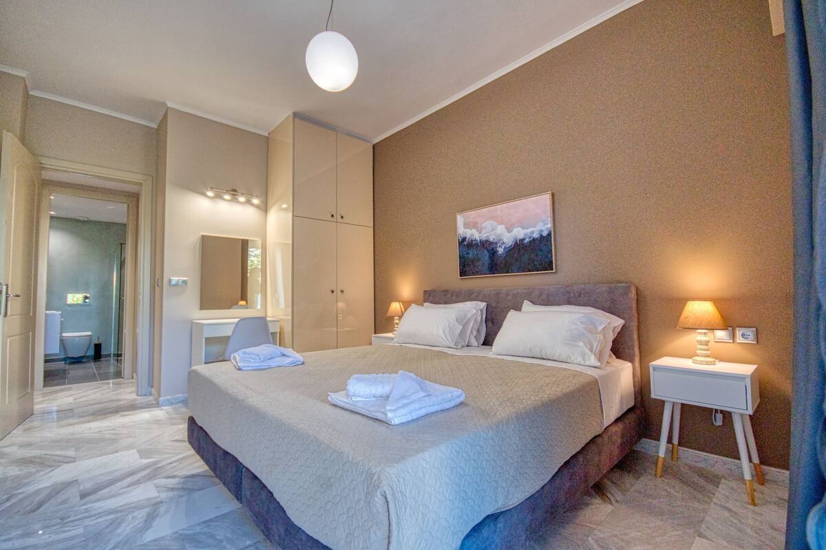 A modern hotel room with a large bed, a bedside table with a lamp, a bathroom with a mirror, and a painting on the wall.