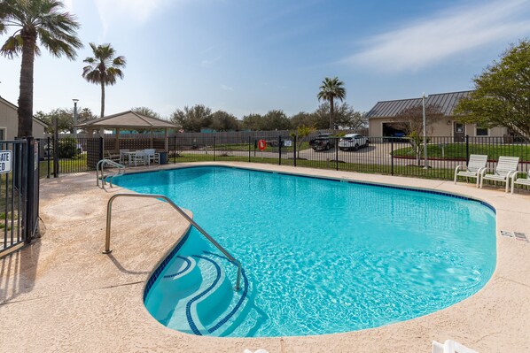 A swimming pool with a curved edge, surrounded by a fence, and a covered seating area with chairs.