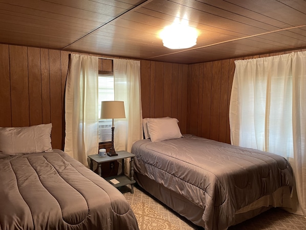 A room with two beds, wooden panel walls, a ceiling light, and a nightstand with a lamp.