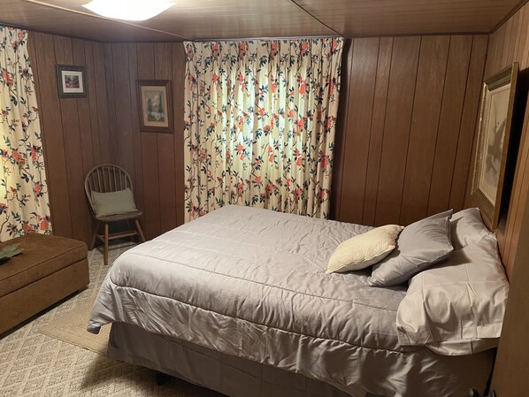 A bedroom with a large bed, floral curtains, a wooden chair, and a framed picture on the wall.