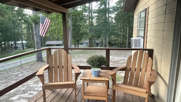 A wooden deck with two Adirondack chairs and a small table, overlooking a wooded area with an American flag. - Cozy Cottage Five (Florien)