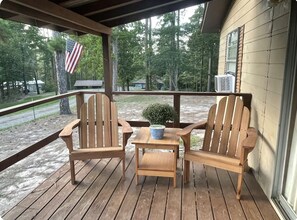 A wooden deck with two Adirondack chairs and a small table, overlooking a wooded area with an American flag.